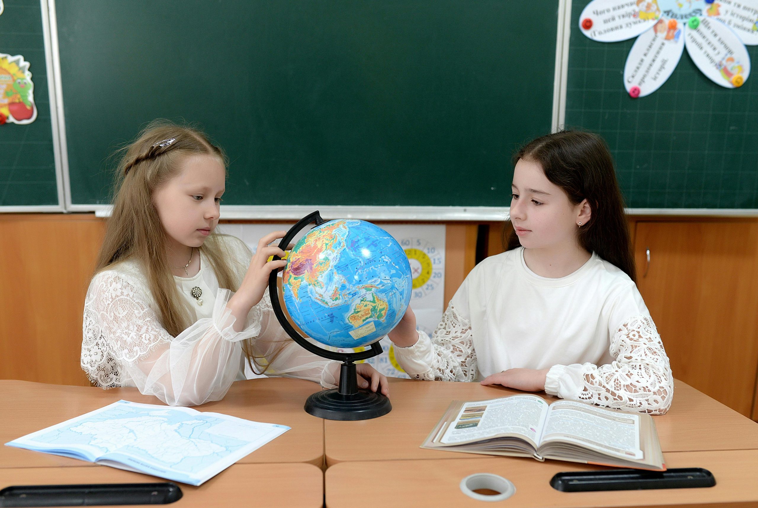Home Two girls studying a globe in a bright classroom environment with educational materials.